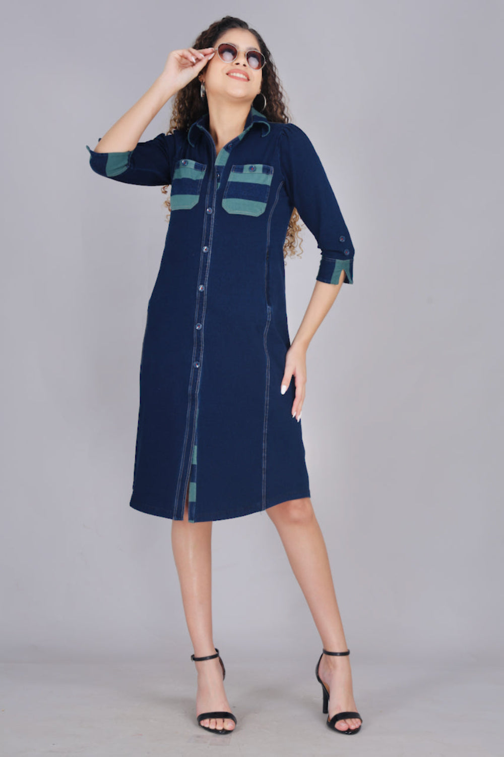 A woman wearing a long denim dress with green and blue striped pockets, button-up front, and two pockets.