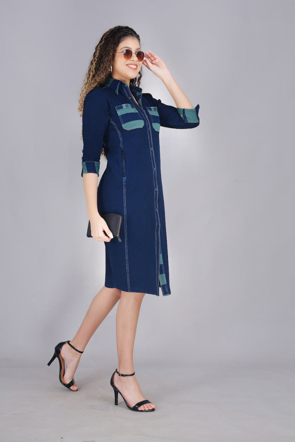 A woman wearing a long denim dress with green and blue striped pockets, button-up front, and two pockets.