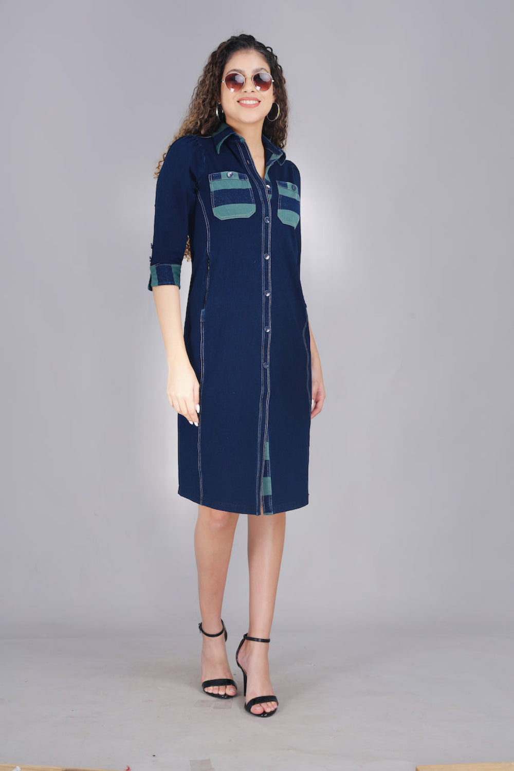 A woman wearing a long denim dress with green and blue striped pockets, button-up front, and two pockets.
