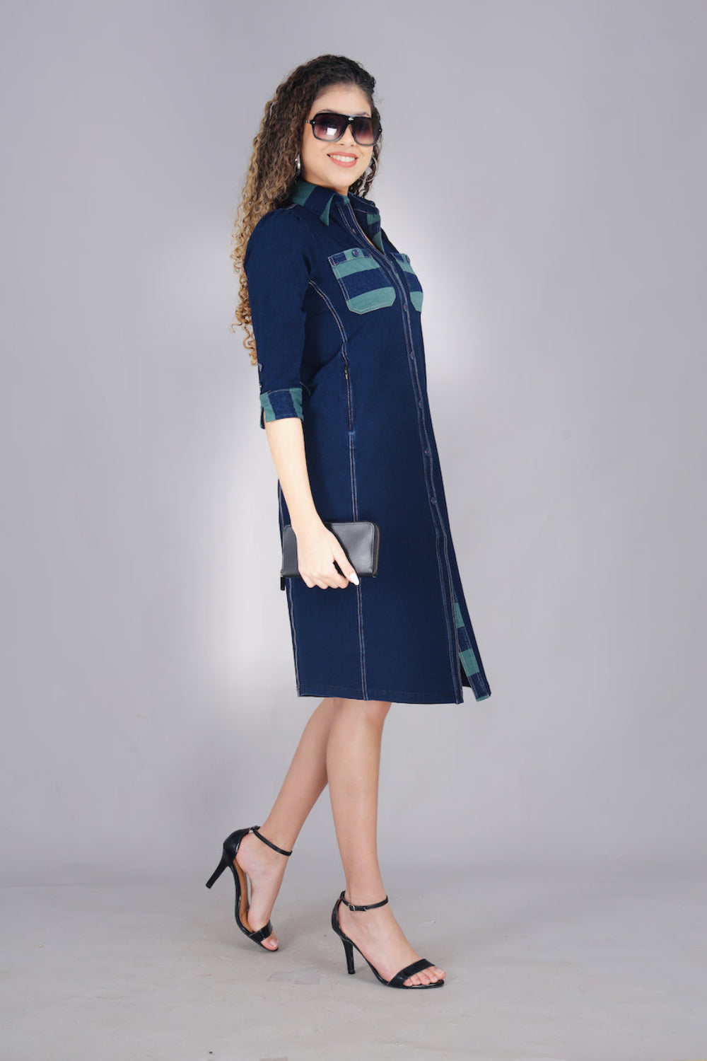 A woman wearing a long denim dress with green and blue striped pockets, button-up front, and two pockets.