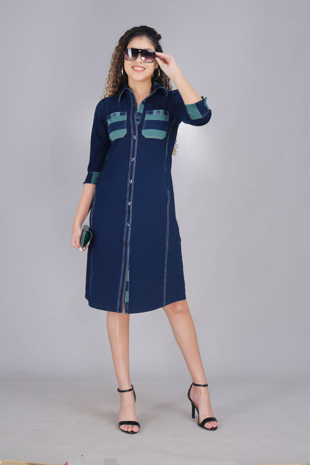 A woman wearing a long denim dress with green and blue striped pockets, button-up front, and two pockets.